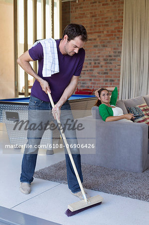 Man cleaning floor with a mop and his wife watching television in the background