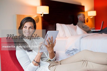 Woman using a digital tablet in a hotel room with her husband using laptop in the background