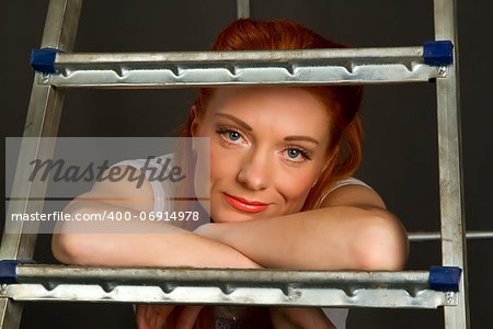 Beautiful young red-haired girl leaning on a ladder on a gray background
