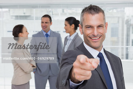 Cheerful businesman in bright office pointing at camera with colleagues on background
