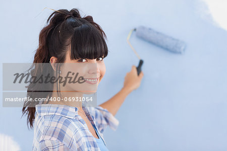 Pretty woman painting wall blue and smiling at camera in new home