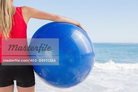 Fit blonde holding exercise ball looking at waves on the beach