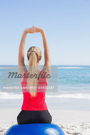 Fit blonde sitting on exercise ball looking at waves stretching arms on the beach