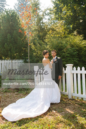 Bride and Groom standing outdoors next to fence on Wedding Day, looking at camera