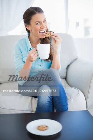 Happy woman holding cup of coffee eating cookie looking at camera