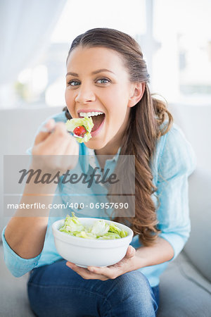 Happy attractive woman eating healthy salad sitting on cosy sofa