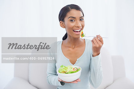 Smiling attractive woman sitting on cosy sofa in bright living room eating salad