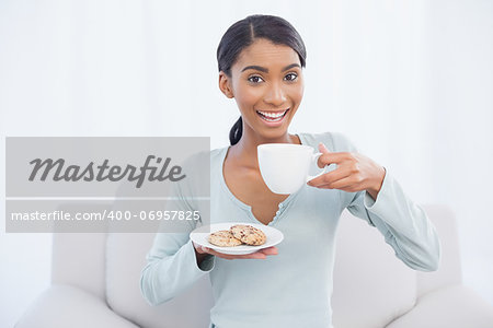 Smiling attractive woman sitting on cosy sofa in bright living room holding coffee and cookies