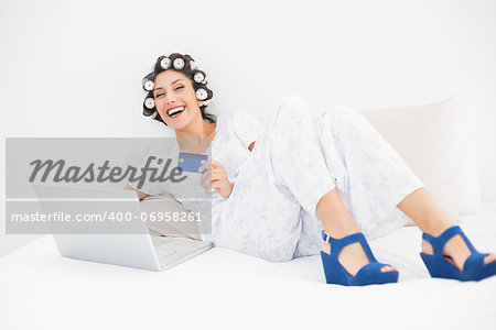 Brunette in hair rollers and wedge shoes using her laptop to shop online in bedroom at home