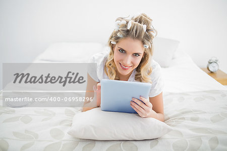 Smiling pretty blonde wearing hair curlers using tablet pc lying on cosy bed