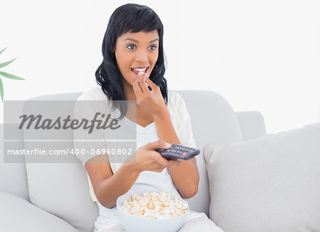 Focused black haired woman in white clothes watching tv while eating popcorn in a living room