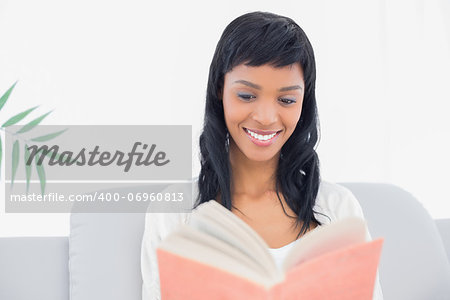 Thoughtful black haired woman in white clothes reading a book in a living room