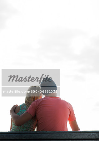 Backview of teenage boy with arm around teenage girl, sitting on bench outdoors, Germany