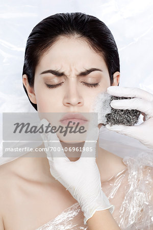 Woman with eyes closed wrapped in plastic, with hands in latex glove scrubbing face with scourng pad, studio shot