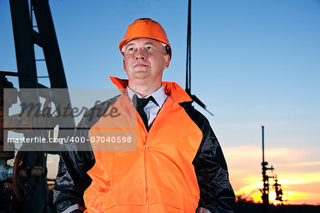 Oil worker in orange uniform and helmet on of background the valves, piping and sunset sky.