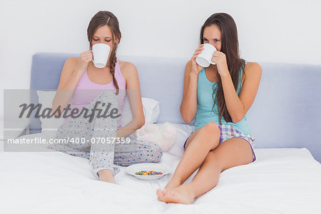 Friends chatting and drinking coffee sitting up in bed wearing pajamas