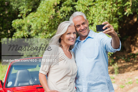 Cheerful mature couple taking pictures of themselves leaning against their cabriolet