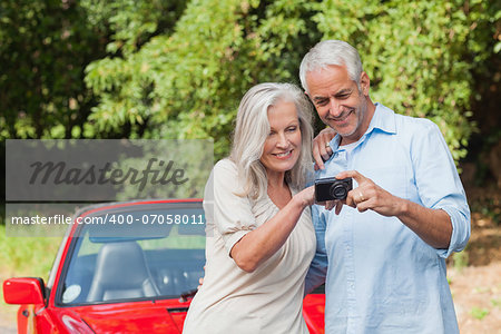 Cheerful mature couple looking at pictures on their camera while leaning against their cabriolet