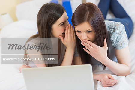 Girl telling a secret to her friend in front of laptop at home in bedroom