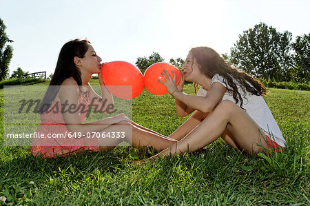 Two young women sitting on grass bowing up red balloons