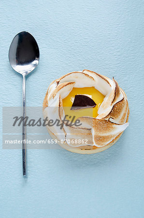 An individual lemon meringue tart and a spoon on a blue surface