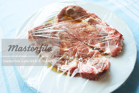 Two pork collar steaks in olive oil marinade on a white plate under cling film