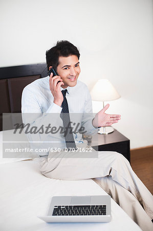 Businessman sitting in front of a laptop on the bed and talking on a mobile phone