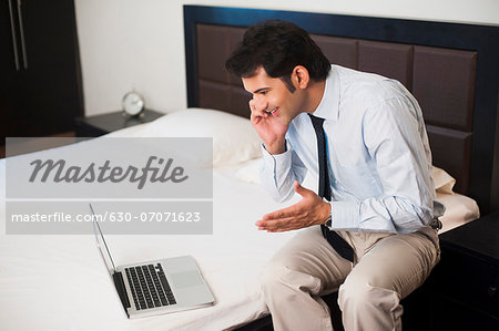 Businessman sitting in front of a laptop on the bed and talking on a mobile phone