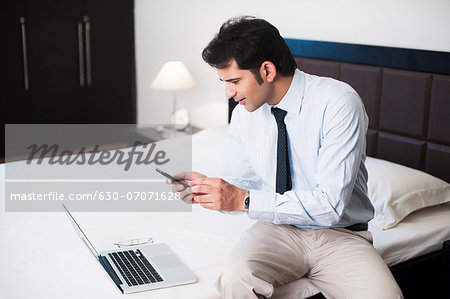 Businessman sitting in front of a laptop on the bed and reading text message on a mobile phone