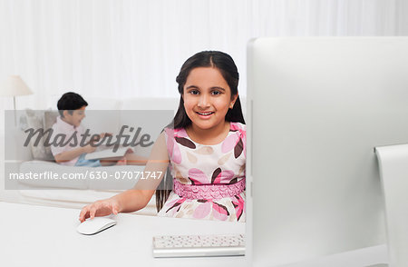 Girl using a computer with her brother reading a book in the background