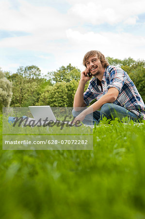 Young man using laptop and cell phone in meadow