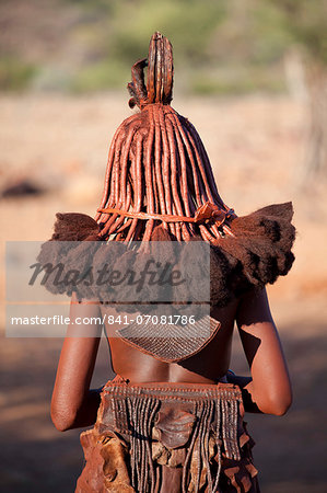 Rear view of young Himba woman showing traditional leather clothing and jewellery, hair braiding and skin covered in Otjize, a mixture of butterfat and ochre, Kunene Region (formerly Kaokoland) in the far north of Namibia
