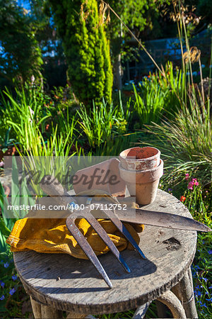 Gardening tools utensils, gloves and flower pots resting on a stool in a green garden with a greenhouse out of focus in the background