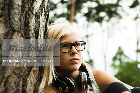 Close-up portrait of girl wearing eyeglasses, sitting next to tree in park, with headphones around neck, Germany Close-up portrait of girl wearing eyeglasses, sitting next to tree in park, with headphones around neck, Germany
