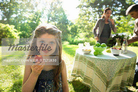 A Young Girl Holding A Large Fresh Organically Produced Strawberry Fruit. Two Adults Beside A Round Table.