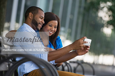 Summer. A Couple Sitting On A Bench, Taking A Selfy Photograph.