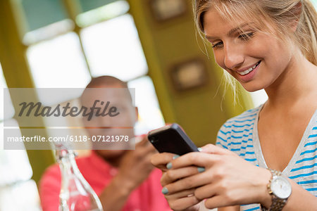 A Couple In A City Coffee Shop. A Woman Sitting Down Checking A Smart Phone. A Man In The Background.