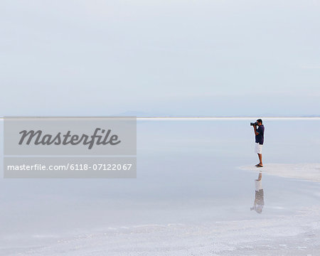 A Man Standing On The Edge Of The Flooded Bonneville Salt Flats, Taking A Photograph At Dusk.