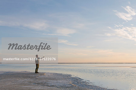 A Man Standing At Edge Of The Flooded Bonneville Salt Flats At Dusk, Taking A Photograph With A Tablet Device.
