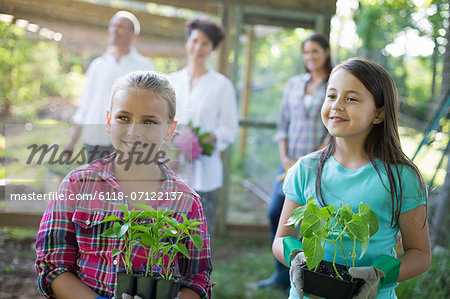 Organic Farm. Summer Party. Two Girls Sitting Holding Young Plants, With A Mature Couple And A Young Woman Looking On.