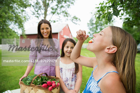 Family Party. Two Young Girls Standing By A Table, One Eating A Fresh Ripe Cherry. A Young Woman Carrying A Bowl Of Salad.