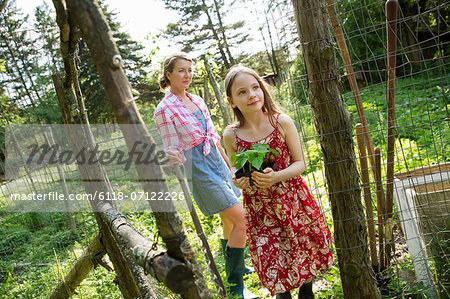 A Woman And A Child Standing At The Open Gate Of A Fenced Enclosure. A Girl Holding A Small Plant In A Pot.