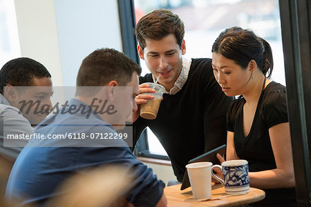A Group Of People Sitting Around A Table In A Coffee Shop. Looking At The Screen Of A Digital Tablet. Three Men And A Woman.
