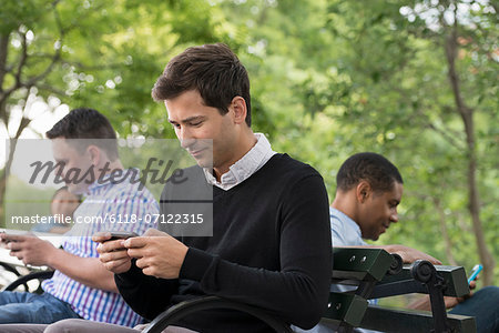 Summer In The City. One Woman And Three Men Sitting In The Park, Each On Their Own Phone Or Using A Tablet.