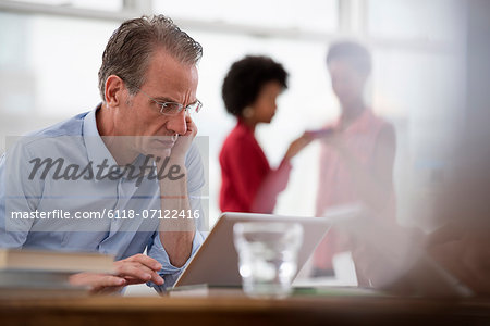 Office Life. A Man Seated At A Computer Laptop Leaning On One Arm, And Two Women In The Background.