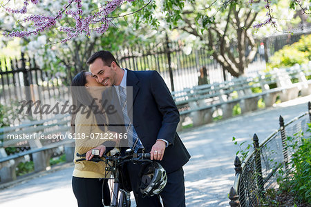A Man In A Business Suit Astride A Bicycle. A Woman Kissing Him Goodbye.