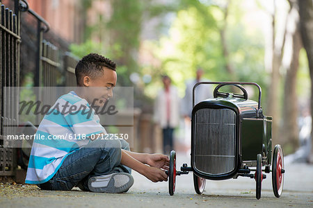 A Young Boy Playing With A Old Fashioned Toy Car On Wheels On A City Street.