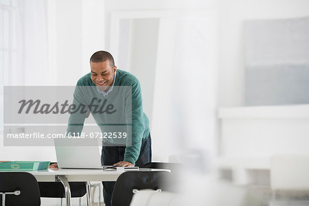 Business. A Man Standing Over A Desk, Leaning Down To Use A Laptop Computer.