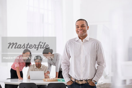 Business. A Man Standing With His Hands In His Pockets, And Three People Leaning Over A Desk Behind Him.