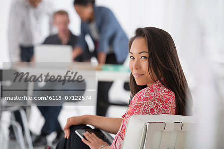 Business. A Woman Sitting With A Digital Tablet On Her Lap, Turning To Look Over Her Shoulder. Three People At A Desk.